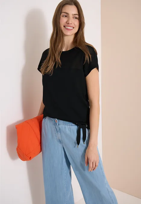 Frau trägt schwarzes T-Shirt, hellblaue Jeanshose und hält orangefarbene Clutch vor neutralem Hintergrund.