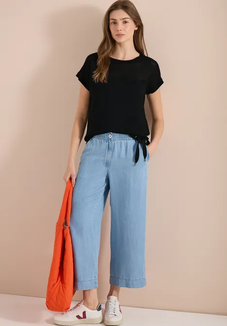 Frau mit schwarzem T-Shirt, weiten hellblauen Hosen, weißen Sneakers und orangener Tasche.