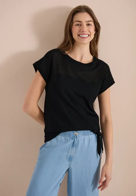 Schlanke Frau trägt schwarzes T-Shirt und helle Jeans, steht vor hellem Hintergrund.
