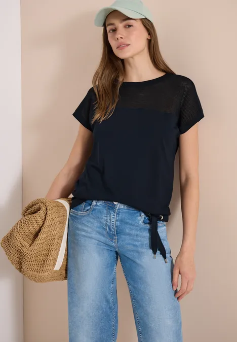 Vrouw in spijkerbroek, donkerblauw T-shirt en pet met strooien tas voor lichte muur