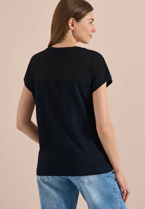 Vrouw van achter gefotografeerd in donkerblauw T-shirt met contrastinzet en lichtblauwe jeans tegen beige achtergrond.