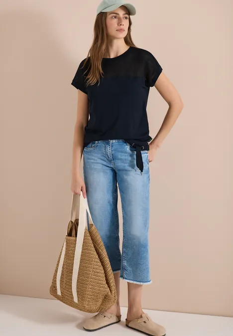 Vrouw in zwart t-shirt, blauwe spijkerbroek, strozak en beige slippers voor een neutrale achtergrond.