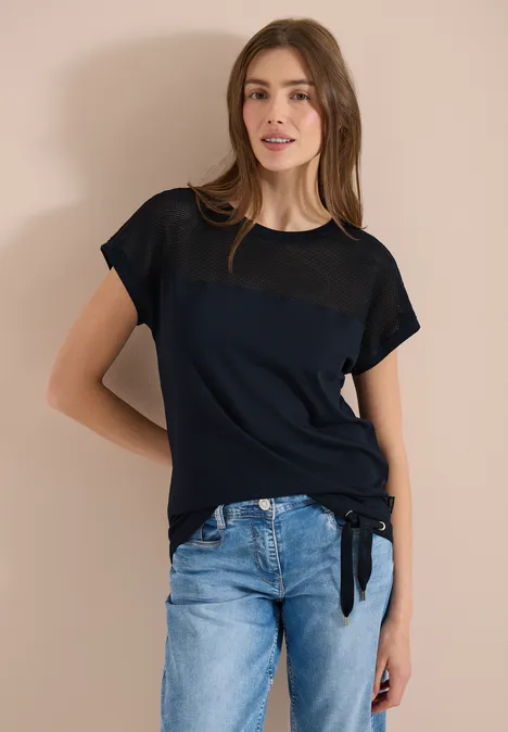 Een persoon in een zwart T-shirt met transparant detail en blauwe spijkerbroek, staand voor een beige muur.