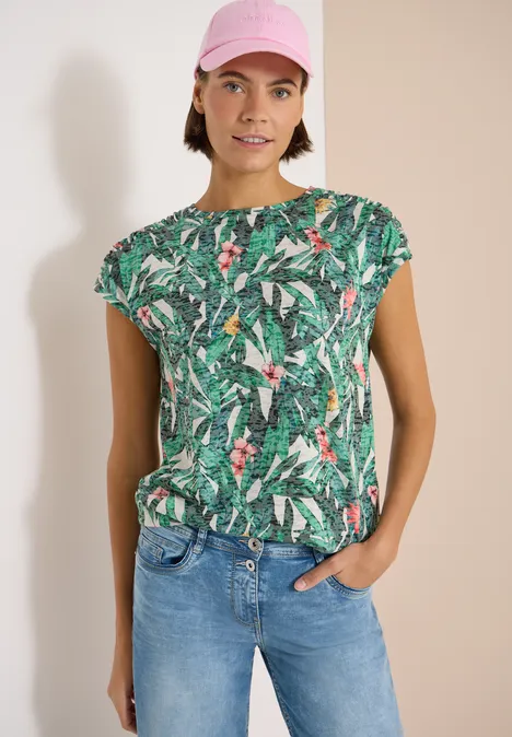 Eine Person trägt ein grünes Shirt mit tropischem Blattmuster und Jeans, steht vor hellem Hintergrund.