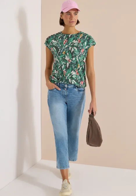 Frau in grünem Tropenprint-T-Shirt, hellblaue cropped Jeans, weiße Sneakers und braune Tasche.