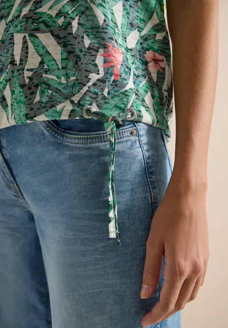 Person trägt blaue Jeans und grüne, gemusterte Bluse mit seitlichem Bindeband