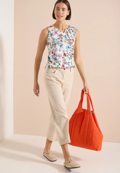 Vrouw in gebloemd topje, beige cropped broek en loafers, met grote rode tas tegen neutrale achtergrond.