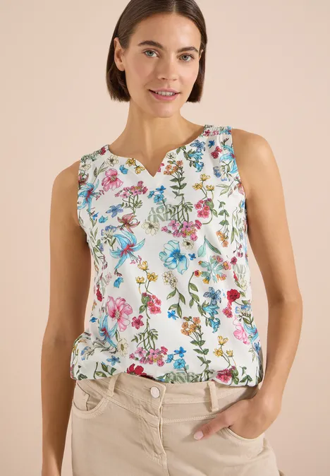 Een vrouw draagt een mouwloos wit topje met kleurrijke bloemenprint en staat ontspannen met hand in de zak.