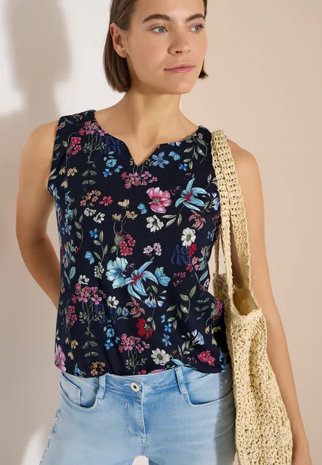 Top mit Split Neck und Raffung universal blue Frau trägt ärmelloses, dunkelblaues Oberteil mit buntem Blumenmuster und eine Stroh-Umhängetasche.