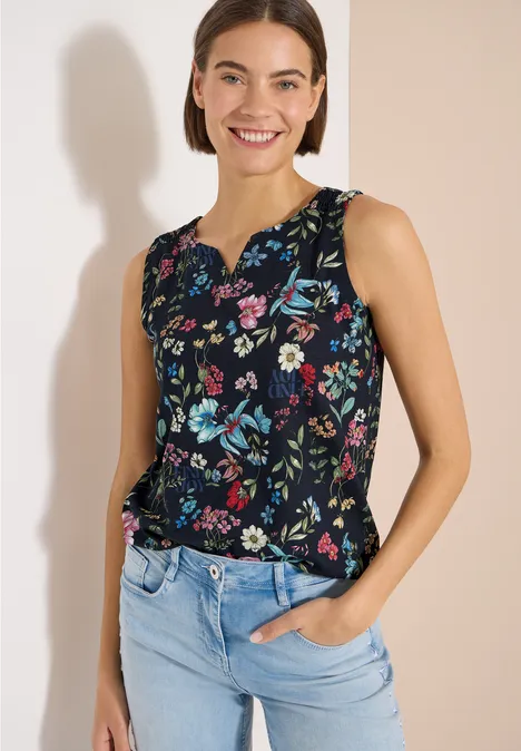 Top mit Split Neck und Raffung universal blue Frau in ärmelloser, dunkelblauer Bluse mit Blumenmuster und hellblauer Jeans vor hellem Hintergrund.