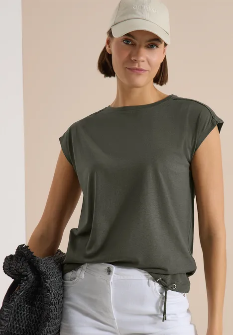 T-Shirt mit Crochet-Details khaki Frau in dunkelgrünem Shirt und hellen Jeans, hält einen schwarzen Pullover vor neutralem Hintergrund.