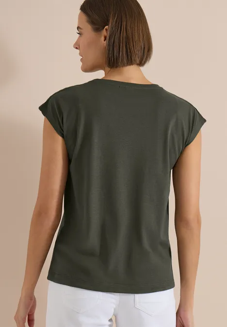 T-Shirt mit Crochet-Details khaki Frau von hinten in dunkelgrünem T-Shirt mit umgeschlagenen Ärmeln und weißer Hose vor beigem Hintergrund.