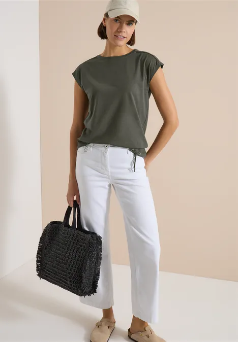 T-Shirt mit Crochet-Details khaki Frau in khakigrünem T-Shirt, weißen Jeans und flachen Sandalen, trägt eine schwarze geflochtene Tasche vor neutralem Hintergrund.