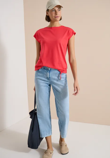 T-Shirt mit Crochet-Details coral Frau in rotem Shirt, hellblauen Jeans und beigen Schuhen mit dunkler Tasche.