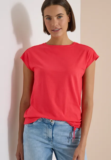 T-Shirt mit Crochet-Details coral Frau in rotem T-Shirt und blauer Jeans steht vor einer Wand, Gesicht unkenntlich.
