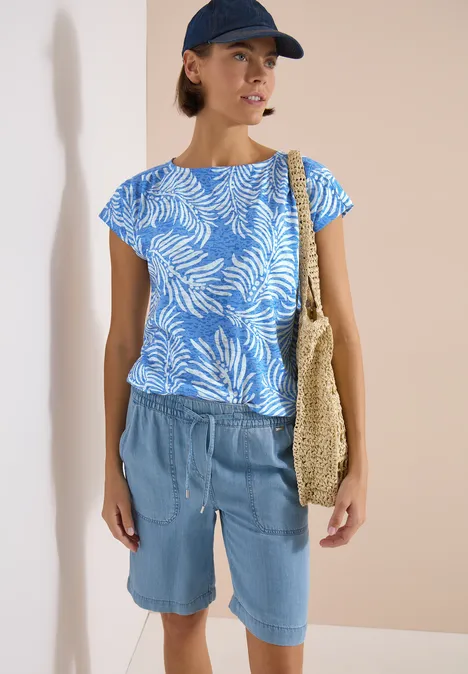 T-shirt à l'effet dévoré null Person mit blau-weiß gemustertem Shirt, Jeans-Shorts, Strohtasche und Cap vor beigem Hintergrund.