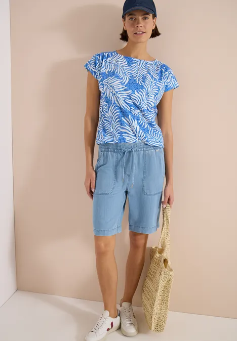 T-shirt à l'effet dévoré null Frau in blau-weißem Blattmuster-Top, lässigen Jeans-Bermudas, weißen Sneakern und Strohtasche vor beigem Hintergrund.