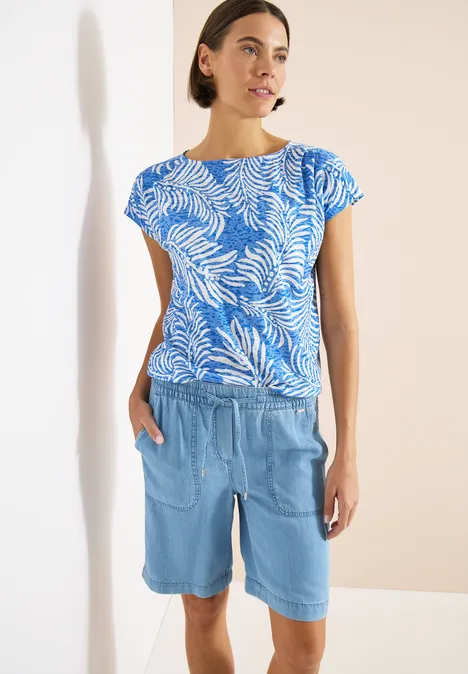 T-shirt à l'effet dévoré null Frau trägt blaues Blattmuster-Shirt und helle Jeans-Bermudashorts vor hellem Hintergrund.