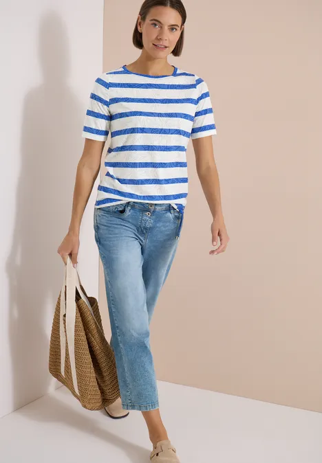 Shirt met burn-out look surf blue Vrouw in blauw-wit gestreept T-shirt, jeans en shopper, lopend voor een lichte muur.