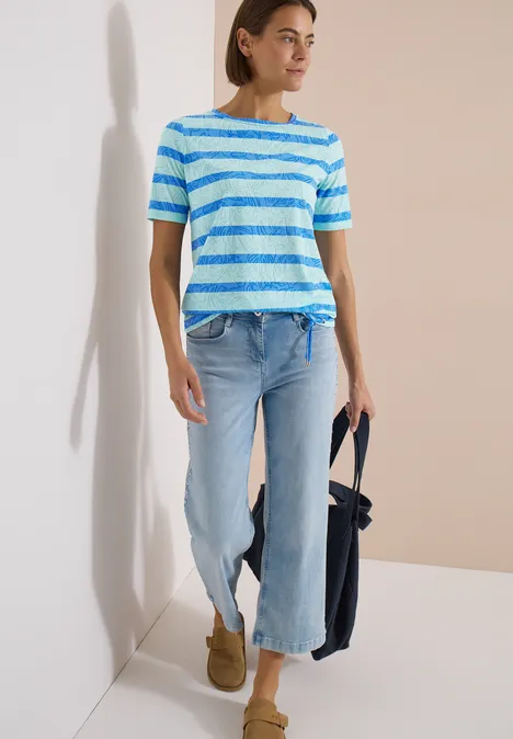 Shirt met burn-out look null Vrouw in blauw-wit gestreept T-shirt, lichtflared jeans, camel laarzen en donkerblauwe tas voor een lichte muur.