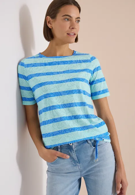 Shirt met burn-out look null Vrouw leunt tegen muur, draagt blauw-groene gestreepte T-shirt en lichte spijkerbroek.