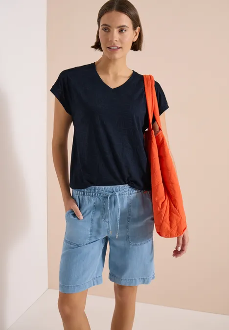 Frau in schwarzem T-Shirt, hellen Jeans-Shorts und rotem Stoffbeutel vor neutralem Hintergrund.