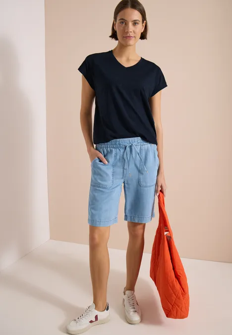 Person in schlichtem Outfit: schwarzes T-Shirt, helle Shorts, weiße Sneaker, orangefarbene Tasche.
