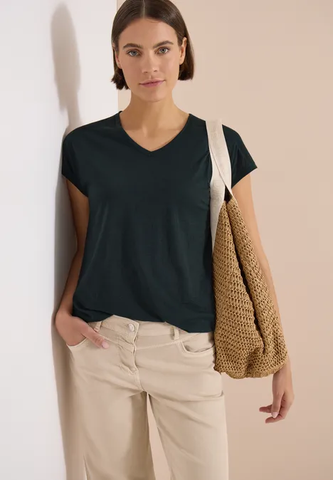 Shirt met ton-sur-ton burn-outpatroon palm breeze green Vrouw in groen V-hals T-shirt en beige broek met gevlochten tas, staand voor een lichte muur
