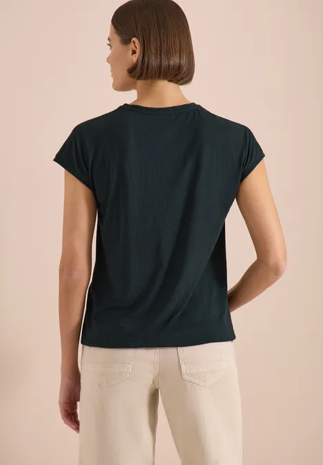 Shirt met ton-sur-ton burn-outpatroon palm breeze green Vrouw met zwarte t-shirt en lichte broek, van achteren gefotografeerd, tegen beige achtergrond.