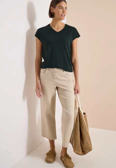 Shirt met ton-sur-ton burn-outpatroon palm breeze green Vrouw in zwart T-shirt met V-hals, beige wijde 3/4-broek, strooien tas en mocassins, lichte achtergrond.