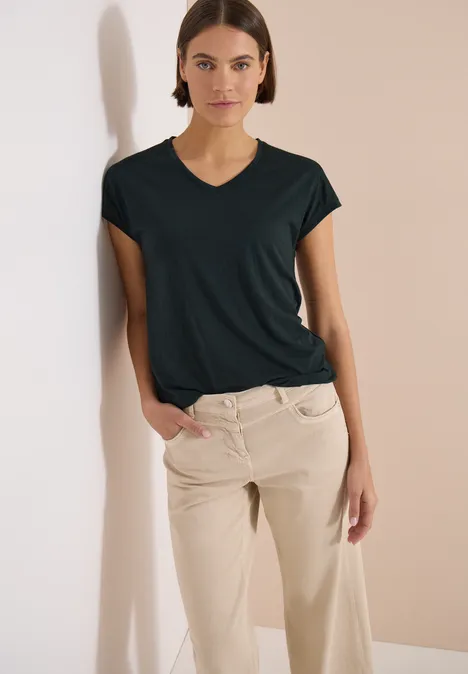 Shirt met ton-sur-ton burn-outpatroon palm breeze green Vrouw leunt ontspannen tegen een lichte muur, draagt zwart shirt en beige broek.