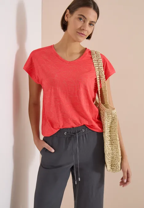 Frau mit rotem T-Shirt, dunklen Stoffhosen und einer Strohtasche vor hellem Hintergrund.