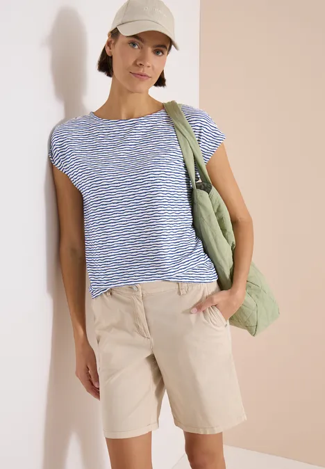 Frau in blau-weiß gestreiftem T-Shirt, beigefarbenen Bermudashorts und grüner Umhängetasche vor neutralem Hintergrund.