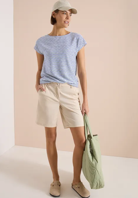 Frau in gestreiftem T-Shirt, beige Shorts und grüner Tasche vor neutralem Hintergrund.