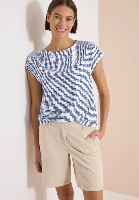 Frau trägt blau-weiß gestreiftes T-Shirt und beige Shorts, Hand in Tasche.