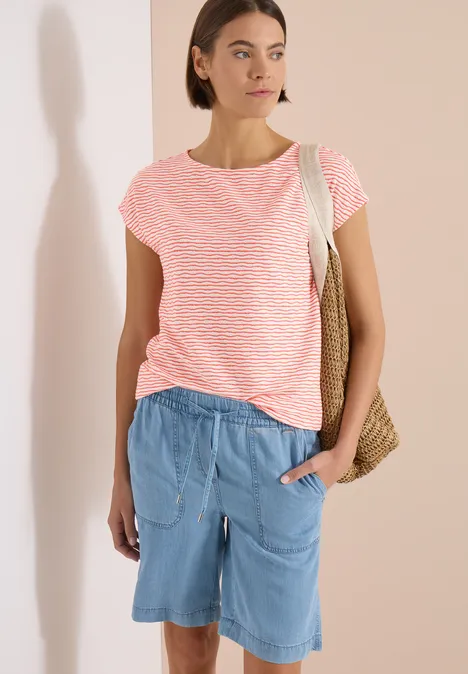 Frau in rosa gestreiftem T-Shirt und blauen Shorts, vor hellem Hintergrund stehend, mit Korbtasche über der Schulter.