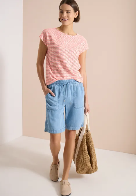 Frau in rosa T-Shirt, hellblauen Jeans-Shorts, beigen Sneakern und Korbtasche vor neutralem Hintergrund.