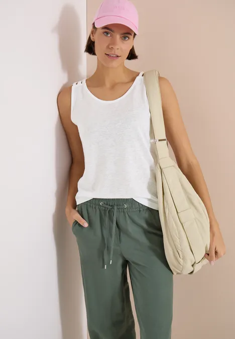 Top avec col en V arrondi et détail tressé White Personne appuyée contre un mur, portant un débardeur blanc, un pantalon vert et un grand sac beige.