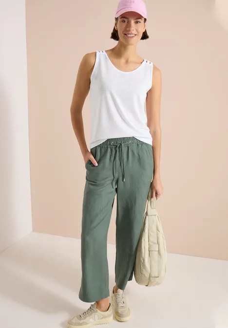 Top avec col en V arrondi et détail tressé White Femme en débardeur blanc, pantalon cargo vert ample, baskets blanches, tenant un sac en tissu beige clair.
