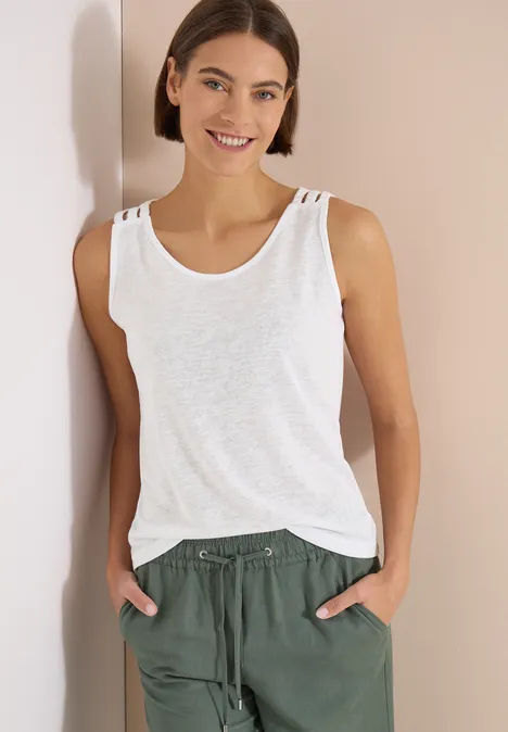 Top avec col en V arrondi et détail tressé White Femme en débardeur blanc et pantalon vert, mains dans les poches, appuyée contre un mur.