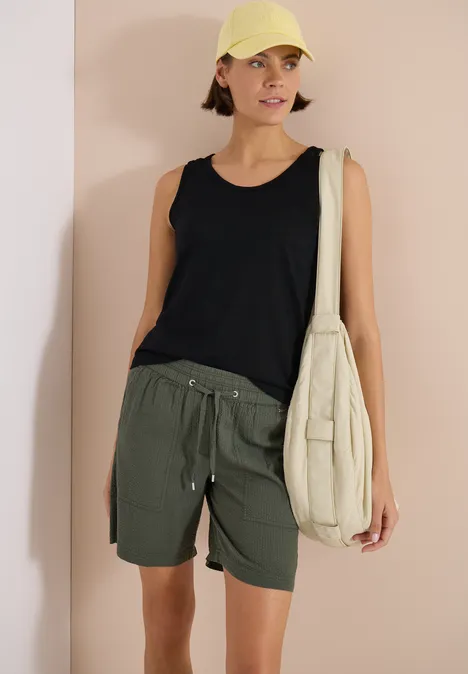 Top met ronde V-hals en gevlochten detail Black Vrouw in zwart hemdje, olijfgroene short en beige schoudertas voor een roze achtergrond.