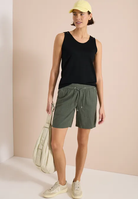 Top met ronde V-hals en gevlochten detail Black Persoon in een zwart tanktop, olijfgroene shorts, beige sneakers en lichtgroene tas voor een lichte achtergrond.