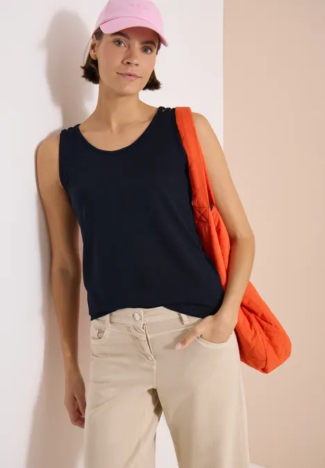 Top met ronde V-hals en gevlochten detail universal blue Vrouw leunt tegen lichte muur, draagt zwarte mouwloze top, beige broek, roze pet en oranje tas.