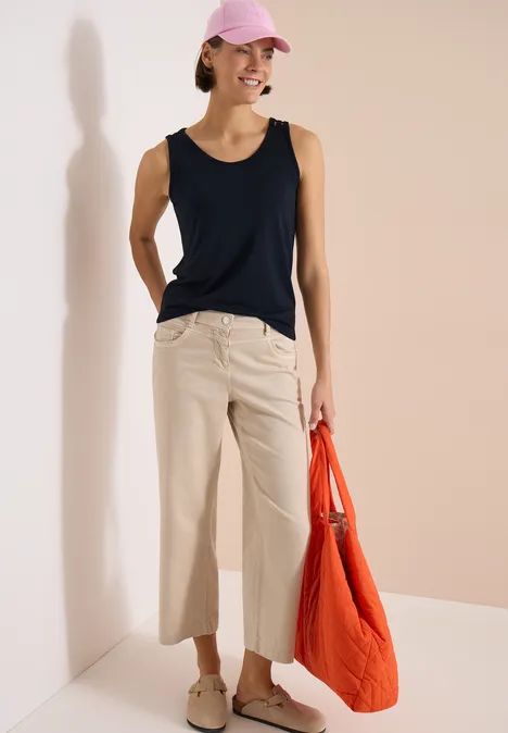 Top met ronde V-hals en gevlochten detail universal blue Vrouw in zwart topje en beige broek met oranje tas tegen lichte achtergrond.