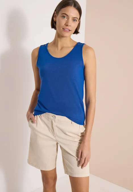 Top avec col en V arrondi et détail tressé surf blue Femme en débardeur bleu et short beige devant un mur crème.