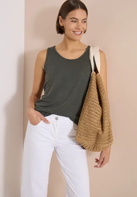 Top met ronde V-hals en gevlochten detail khaki Vrouw in donkergrijs hemd, witte jeans en grote strooien tas, voor een beige achtergrond.