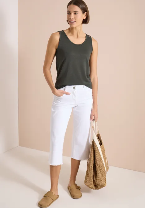 Top met ronde V-hals en gevlochten detail khaki Vrouw in donkergroene top, witte driekwartsbroek, beige slippers en strozak voor een neutrale achtergrond.
