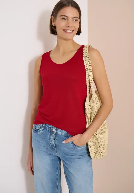 Top avec col en V arrondi et détail tressé tamarillo red Femme en débardeur rouge, jean bleu et sac en paille devant un fond neutre.