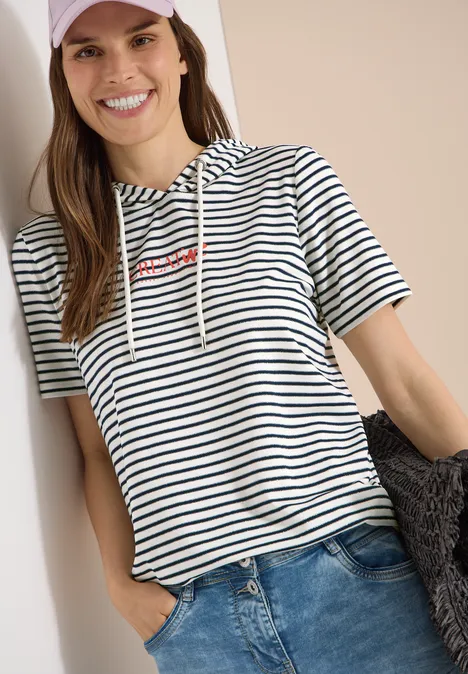 Shirt met korte mouwen, capuchon en glitter vanilla white Vrouw leunt ontspannen tegen een muur, draagt gestreept T-shirt met capuchon en jeans, één hand in de zak.