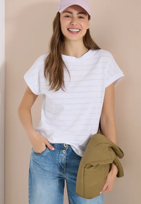 Frau in weißem T-Shirt und blauen Jeans, hält grüne Jacke, vor neutralem Hintergrund.